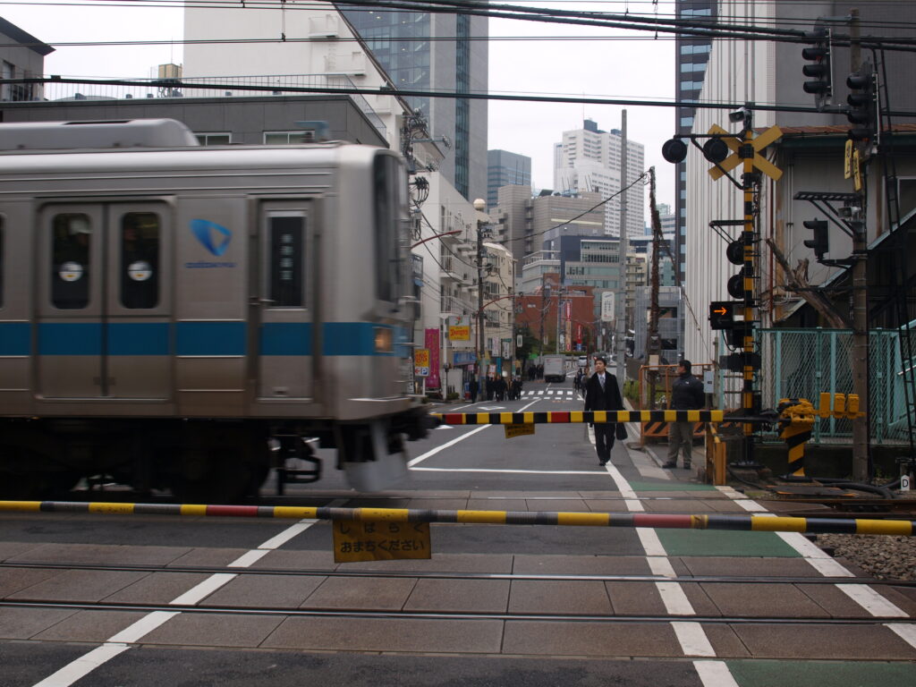 Street photograph of a train passing a level crossing in Tokyo. In the foreground are barricades and tracks; a silver-blue train is moving to the left; pedestrians and city buildings are in the background under a gray sky.
