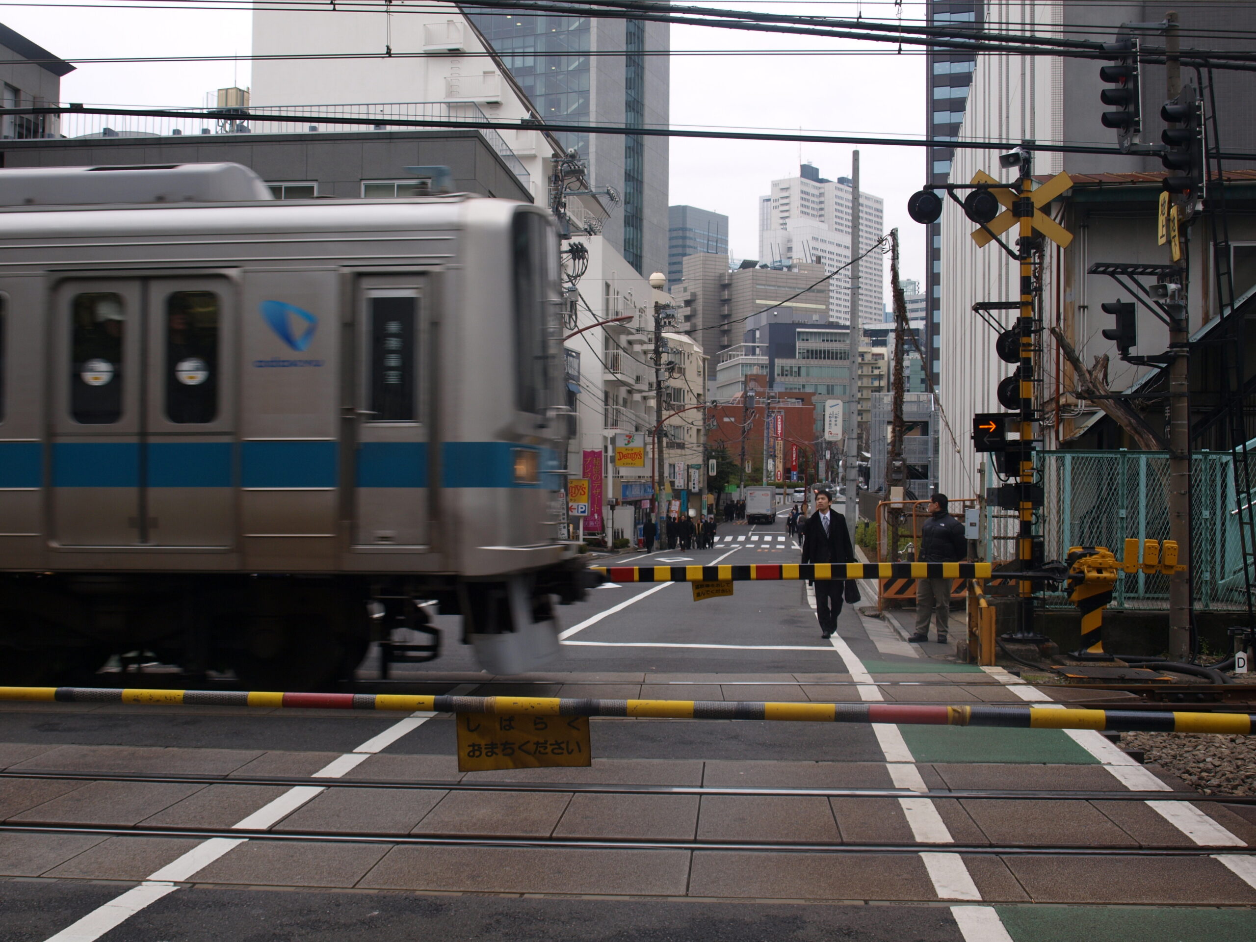 Street photograph of a train passing a level crossing in Tokyo. In the foreground are barricades and tracks; a silver-blue train is moving to the left; pedestrians and city buildings are in the background under a gray sky.