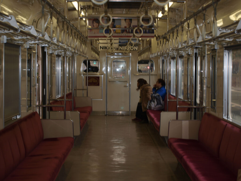 The photo shows the interior of a train in Japan with empty seats on both sides, handrails hanging from the ceiling, and two passengers sitting on the right side. The scene is viewed from the aisle toward the door at one end, with soft, warm colors and reflections in the windows.