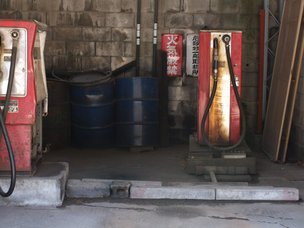 Photograph of a worn-out garage or gas station interior featuring two red pumps, blue oil drums, and a concrete wall in the background. The scene is rendered in subdued, earthy, neutral tones and shows the wear and patina on the machinery.