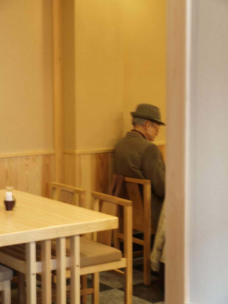 A portrait photograph of an elderly person wearing a hat, sitting alone on a wooden chair along the wall in a warmly lit café or restaurant; the foreground shows an empty wooden table and chairs; the image is soft and slightly blurred.