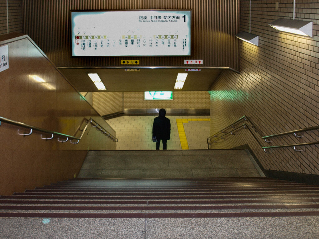 A digital photograph of a subway station taken from the top of a staircase; a single person is standing with their back to the camera at the bottom of the image, surrounded by beige tiles, metal handrails, signage, and a yellow guide line on the floor.