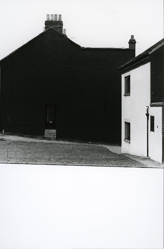 Black-and-white photograph of two buildings as seen from a lawn; a dark, windowless facade on the left with a small door and chimney tops, and a lighter-colored gable with windows on the right. The lower part of the image is a large white area.