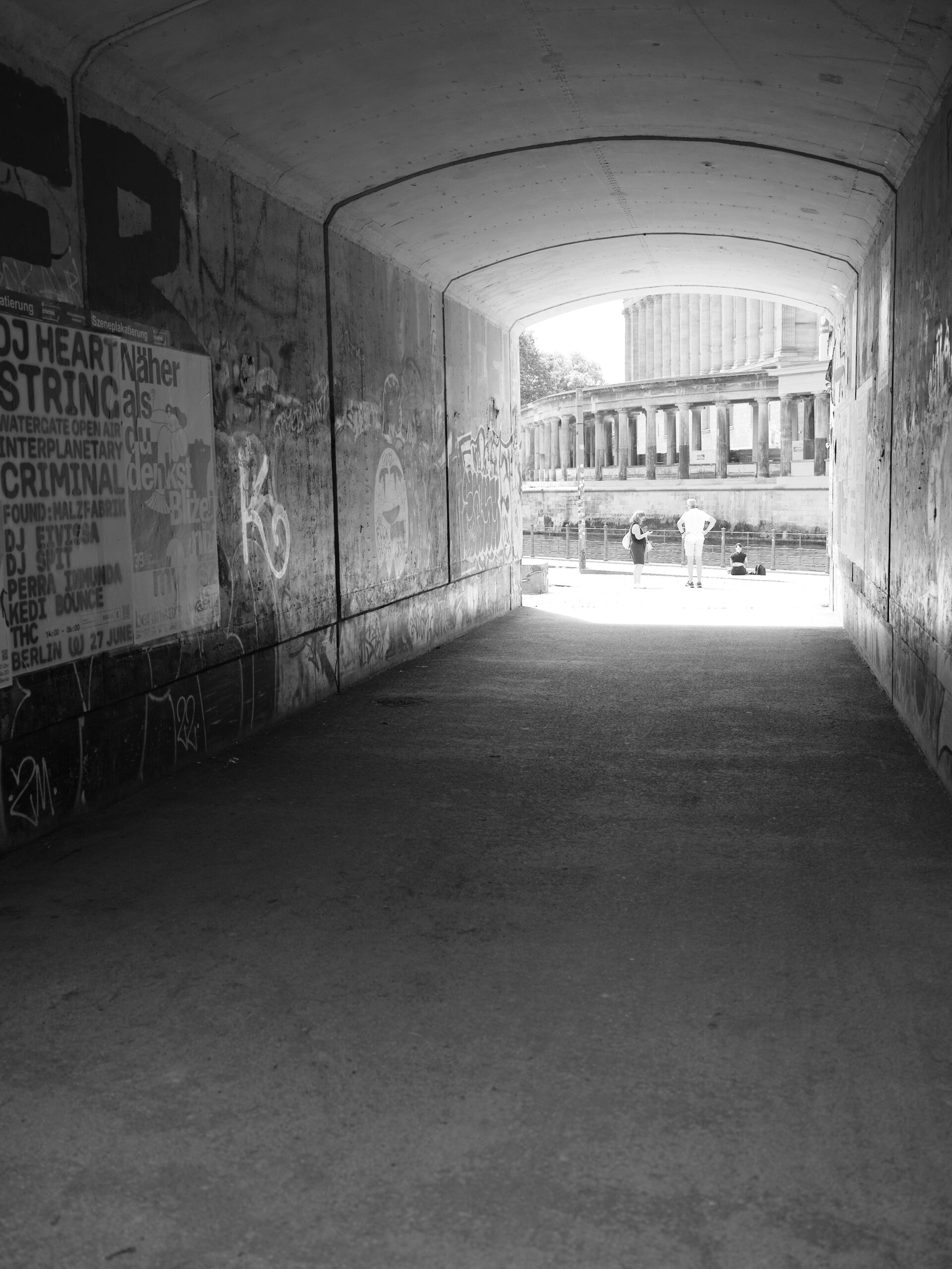 A black-and-white photograph taken from a dark tunnel or passageway toward a bright, open space by a river; graffiti on the tunnel walls; and three people in the clearing in front of a classical columned building overlooking the water.