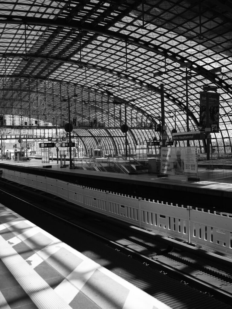 Black-and-white photograph of a station concourse with a large arched glass-and-steel structure, empty platforms, signage, and tracks in the foreground; strong shadows and graphic patterns cast by the roof structure.