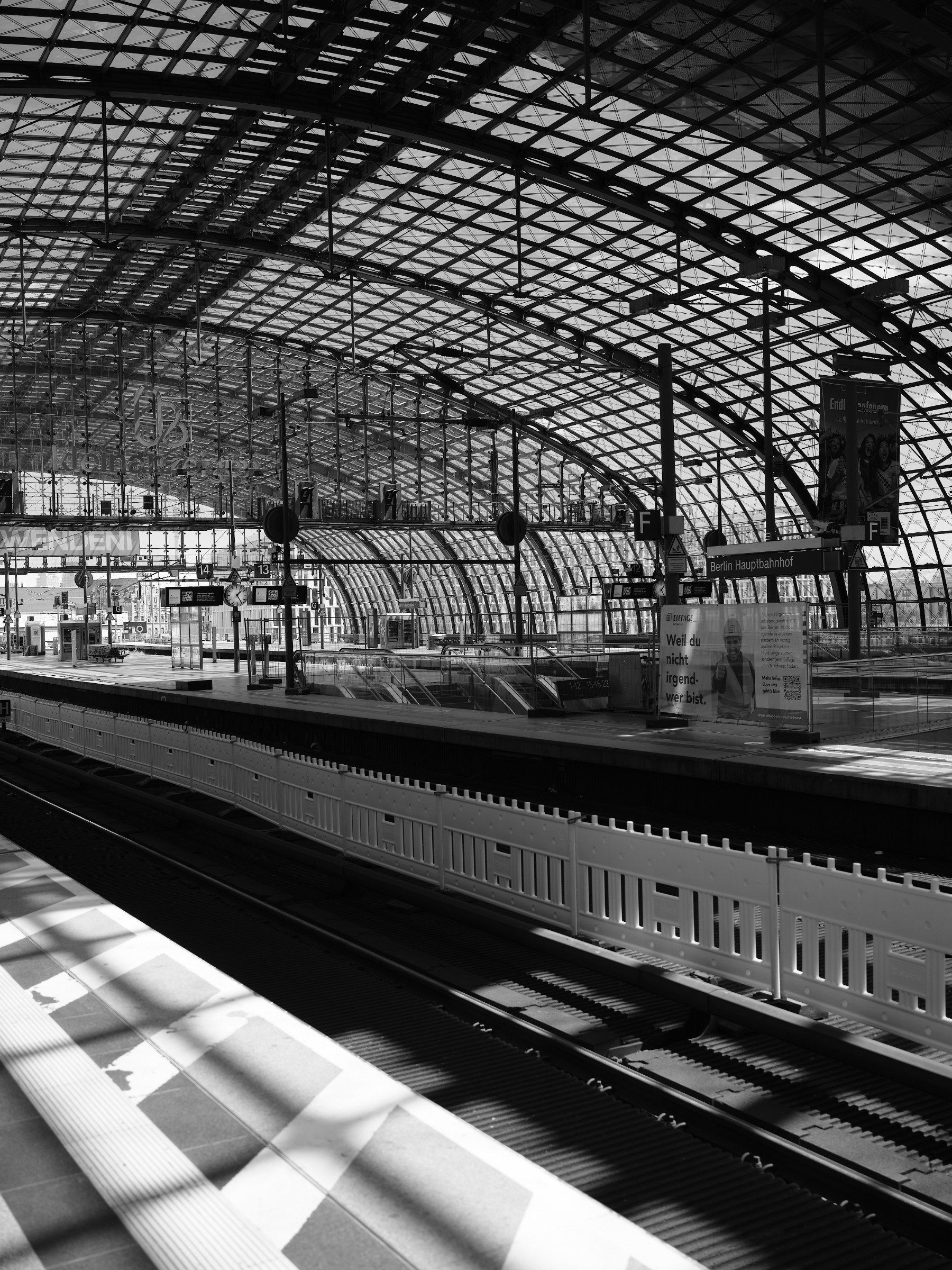 Black-and-white photograph of a station concourse with a large arched glass-and-steel structure, empty platforms, signage, and tracks in the foreground; strong shadows and graphic patterns cast by the roof structure.