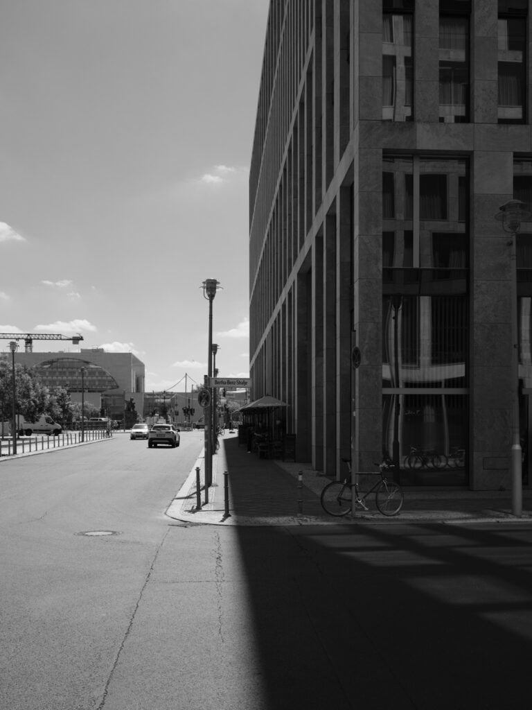 Black-and-white photograph of a city street in Berlin with a tall office building on the right; a sharp shadow divides the street into light and dark; a bicycle parked by the sidewalk, cars in the background, and a sense of open perspective toward the horizon.