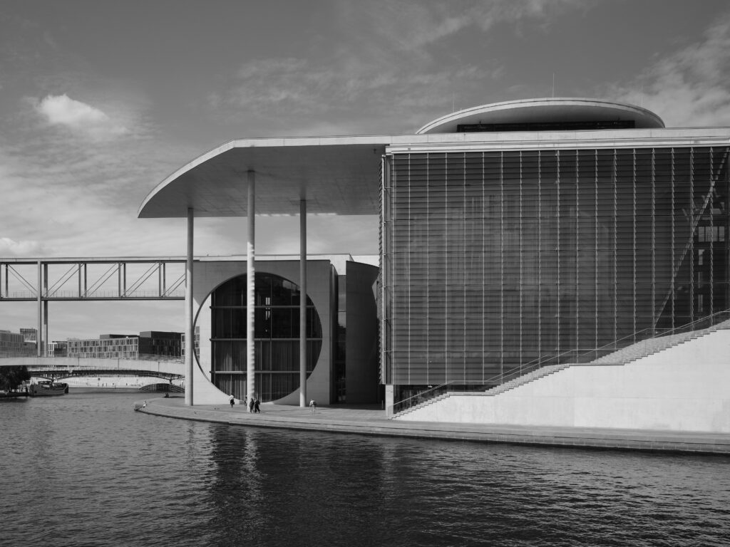 Black-and-white photograph of modern architecture along a riverbank: a large building with round and rectangular shapes, columns, and an overhanging roof structure. Stairs leading down to the water’s edge, a walkway in the background, and people walking along the promenade. Reflections in the water and a cloudy sky.