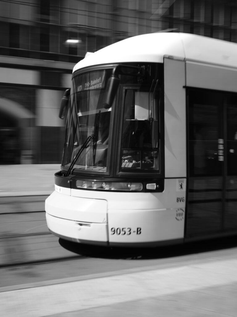 Black-and-white photograph of a close-up of a modern tram in motion on an urban street; the image is partially blurred by motion, with the focus on the front of the tram, where the number 9053-B is visible.