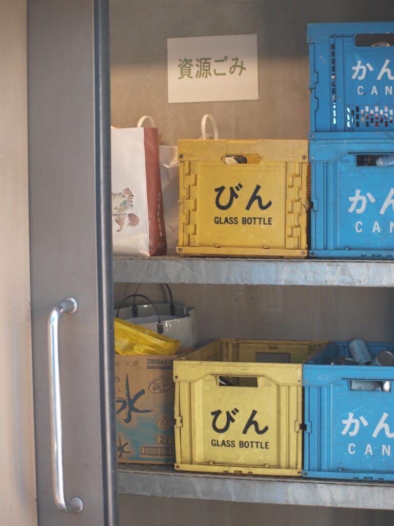 Photograph of a storage shelf with yellow and blue plastic boxes labeled "Glass Bottle" and "Can," bags, and cardboard boxes, seen through a metal frame/door handle on the left side.
