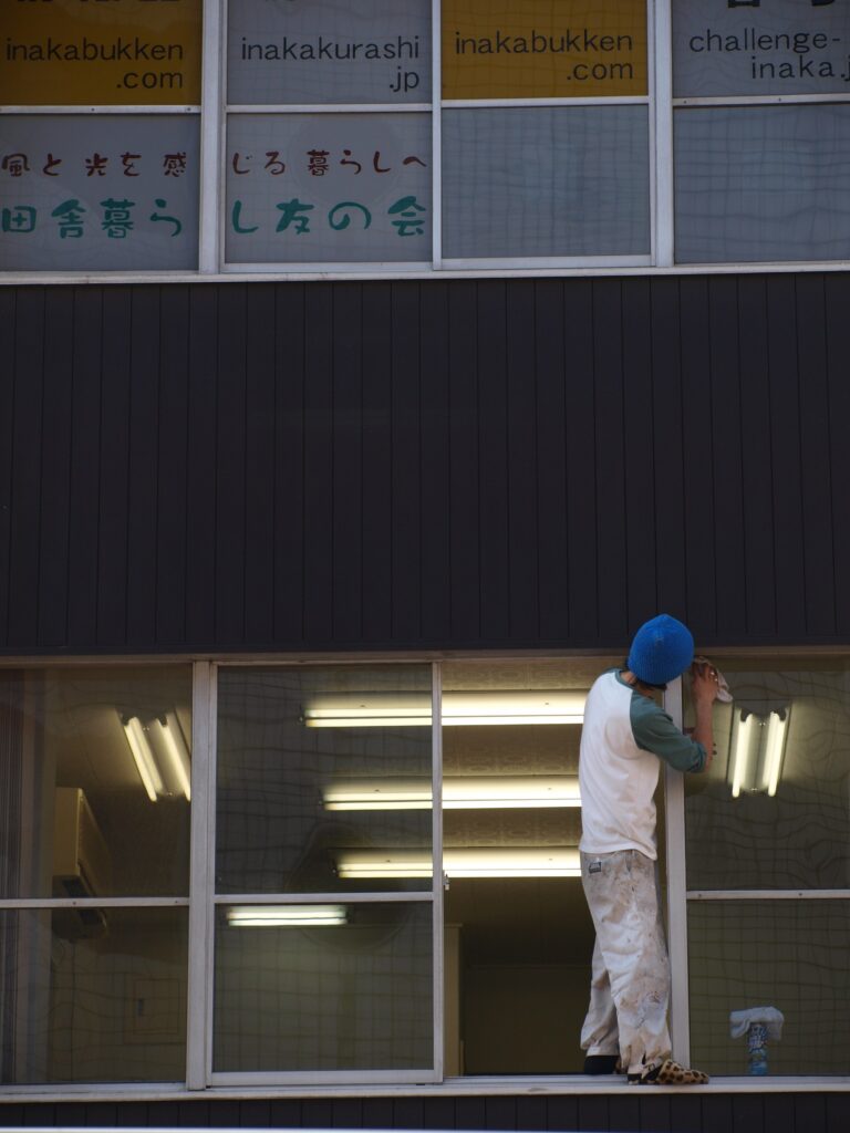 Photograph of a person wearing a blue cap and a white/green sweater working on a window frame in a building with large windows; the upper part shows signs with Japanese text, and fluorescent light from inside is reflected in the glass.