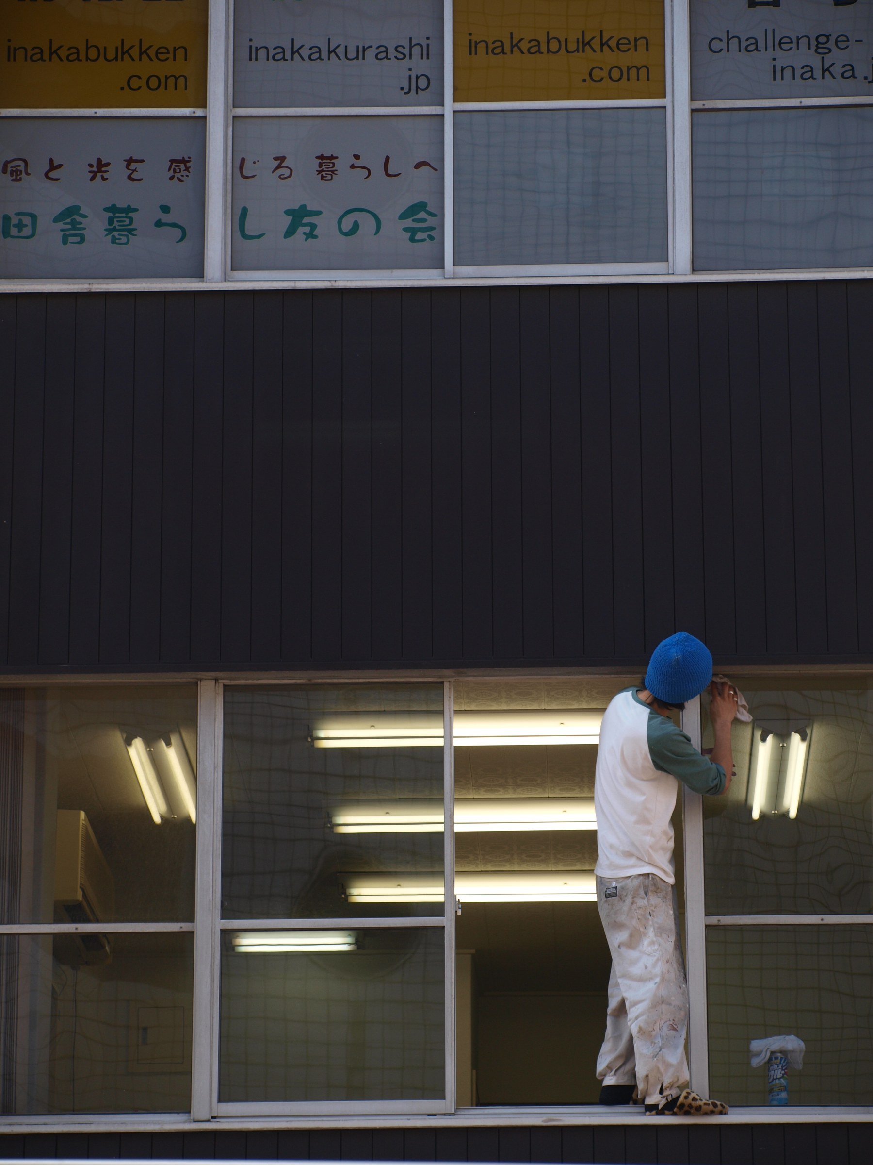 Photograph of a person wearing a blue cap and a white/green sweater working on a window frame in a building with large windows; the upper part shows signs with Japanese text, and fluorescent light from inside is reflected in the glass.