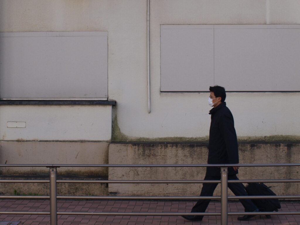 Street photograph of a person wearing a face mask and a coat, walking to the right along a bridge with a metal railing in front of a weathered white wall with boarded-up windows. The person is pulling a suitcase.