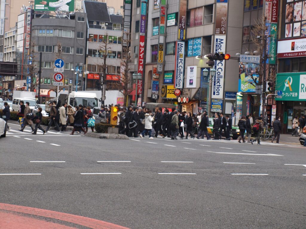 Street photograph of a busy crosswalk in a city with many people crossing and tall storefronts and advertising signs in the background. The street in the foreground is wide and free of cars, while a crowd of people is crossing at a traffic signal in the center of the image.