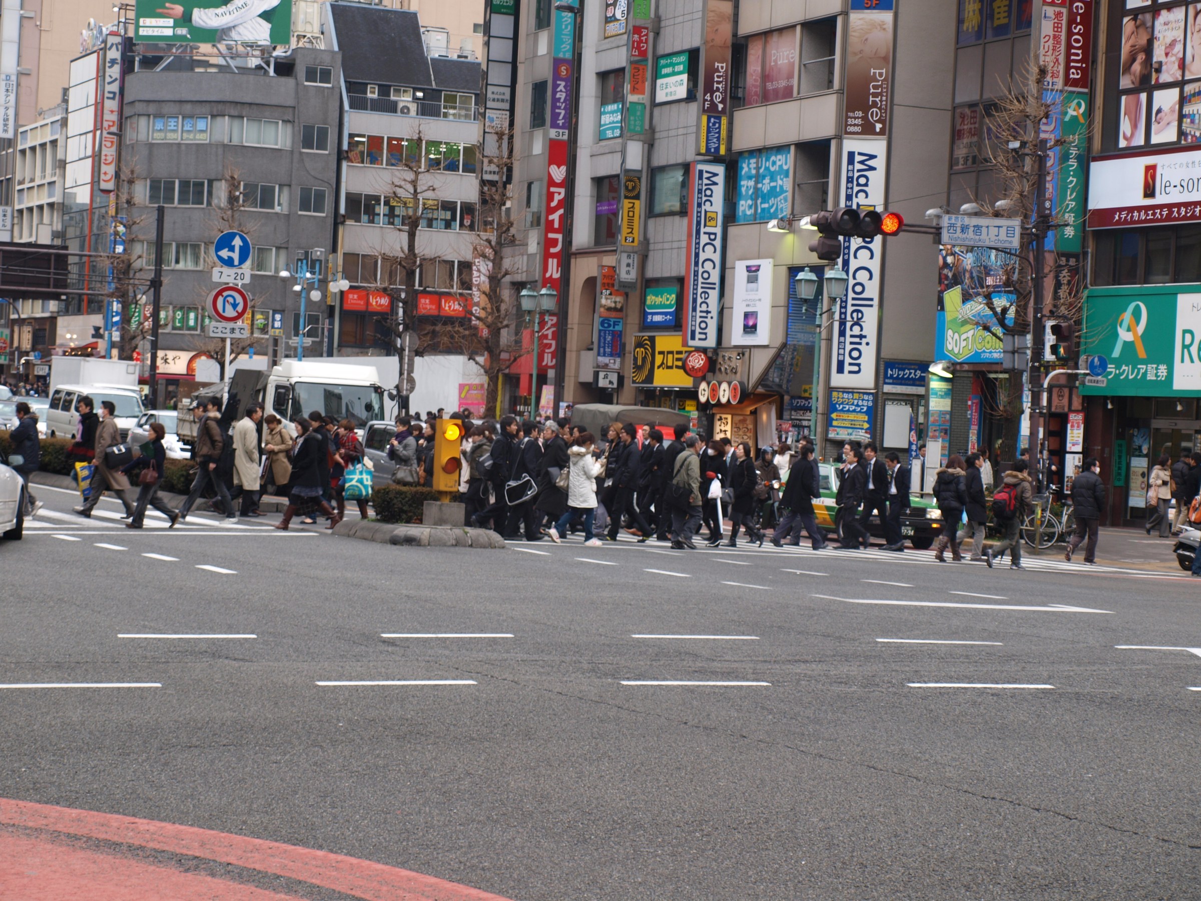 Street photograph of a busy crosswalk in a city with many people crossing and tall storefronts and advertising signs in the background. The street in the foreground is wide and free of cars, while a crowd of people is crossing at a traffic signal in the center of the image.