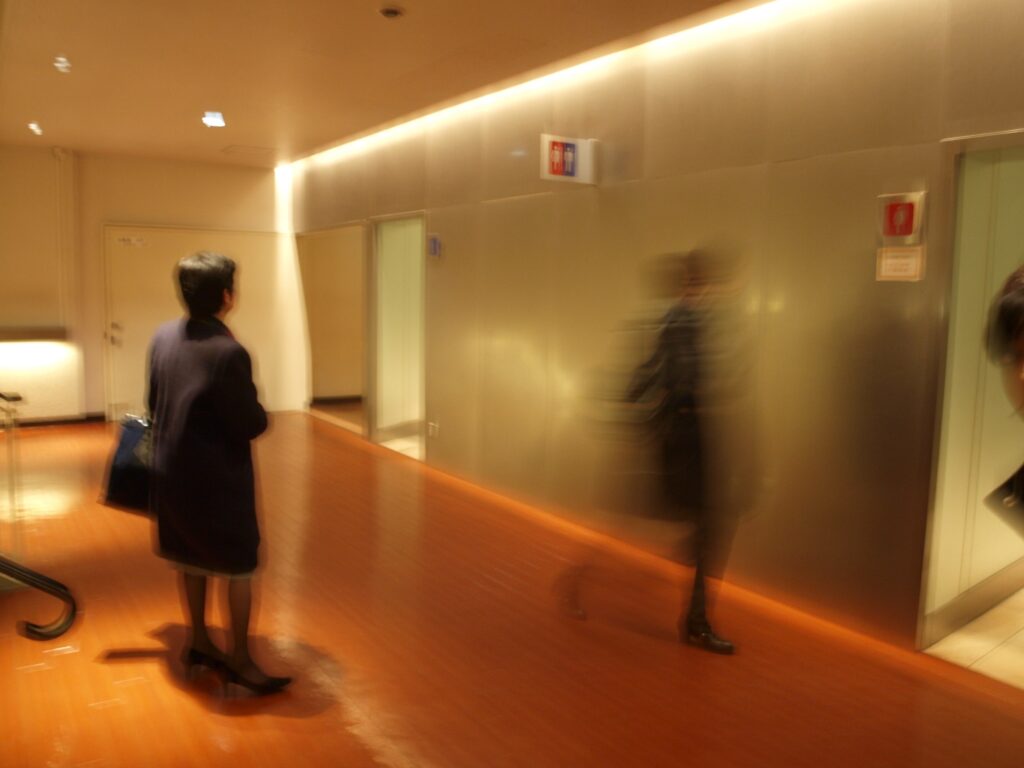 A blurry indoor photograph of people in a hallway with warm lighting and reflective metal walls; one figure is standing and waiting while another is blurred in motion, with a wooden floor and doorways visible.