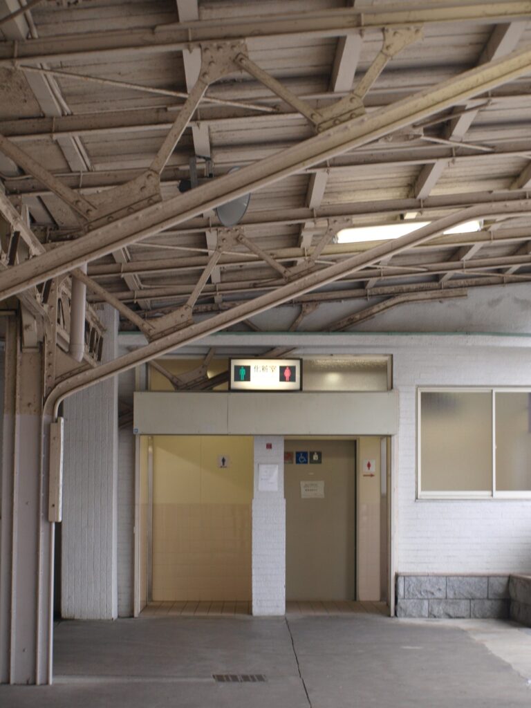 Photograph of a station building or platform area with visible metal roof trusses, an entrance to restrooms with signs, a skylight, and a window on the right; exposed concrete floors and muted neutral colors.